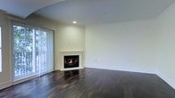 This two-floor property offers a well-appointed living space characterized by a refined yet understated aesthetic. The main living area immediately draws attention with its white-mantled fireplace, which serves as a natural focal point and adds warmth to the room. Dark hardwood flooring runs throughout, providing a rich contrast against the bright, neutral-toned walls and contributing a sense of depth and elegance to the space.

Recessed lighting in the ceiling supplements natural light that filters in through vertical blinds covering a large window or sliding door, hinting at outdoor access and views of surrounding greenery. The smooth, light grey ceiling pairs cleanly with white crown molding and baseboards that frame the room with a classic, polished finish.

The open layout of the living area offers generous space and flexibility for a variety of furniture arrangements. The walls are neutral and move-in ready, providing a blank canvas for personalization. The overall ambiance is calm and inviting, combining timeless design elements such as the fireplace and hardwood floors with a clean, modern simplicity that suits a range of interior styles.