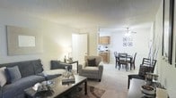 This single-floor residence showcases an open-concept design that seamlessly connects the living room, dining area, and kitchen into one cohesive, flowing space. The living room is carpeted and comfortably furnished with a sofa, armchair, and a glass-top coffee table, all arranged around a decorative area rug. Neutral wall tones and a textured ceiling contribute to a calm, understated ambiance throughout. A ceiling fan with a light fixture overhead serves the dining area, where a dark-framed glass dining table and matching chairs provide a functional yet stylish setting. The kitchen, visible in the background, features warm wood-toned cabinetry and appears equipped with standard appliances, including a range and overhead cabinets for storage. A paneled door near the living area provides access to an adjacent room. Natural and artificial lighting work together to keep the space bright and welcoming. The open layout maximizes the sense of spaciousness, making this residence feel connected and easy to navigate, while the consistent neutral palette and carpeted flooring unify the overall aesthetic.