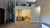 This two-story space makes a striking first impression with its soaring double-height ceilings and open, airy layout. A sleek spiral staircase anchors one side of the main floor, serving as both a functional element and a bold architectural focal point that draws the eye upward. The upper level features an open loft with wrought iron railing, offering a bird's-eye view of the living area below and lending the space a dynamic sense of vertical depth.

On the main floor, a recessed dining nook provides a cozy, defined area for a dining table and chairs, framed by warm neutral walls accented with a contrasting wainscoting-style lower panel. A ceiling fan overhead adds both comfort and a casual, homey touch to this intimate space. The adjoining area offers a glimpse into additional rooms beyond, hinting at a thoughtful and connected floor plan.

Plush carpeting throughout the main level adds warmth and comfort underfoot, complementing the neutral, inviting color palette. The combination of open vertical space, architectural detailing, and well-defined functional zones creates a balanced ambiance that is both practical and visually engaging — ideal for everyday living and entertaining alike.