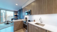 This contemporary single-floor residence showcases a refined modern aesthetic throughout its thoughtfully designed layout. The kitchen is a standout feature, outfitted with warm wood-grain cabinetry extending to the ceiling, crisp white quartz countertops, a sleek glass backsplash, and a full suite of stainless steel appliances including a range, over-the-range microwave, and dishwasher. Light wood flooring carries through the open-concept living and dining area, where floor-to-ceiling windows frame impressive urban skyline views and flood the space with natural light. The living area is designed for comfort and sophistication, featuring textured dark walls, a plush sofa, a cognac leather lounge chair, a compact dining nook, and curated artwork. The bedroom continues the contemporary theme with light hardwood floors, a dark accent wall adorned with framed artwork, a walk-in closet, and a large window offering sweeping city views. The full bathroom is finished with large-format neutral floor tiles, wood-tone vanity cabinetry, a white quartz countertop, and a full-size soaking tub with shower. Throughout the residence, the design balances warm natural materials with cool, modern finishes to create a cohesive and inviting atmosphere.
