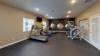 This fitness center is a thoughtfully designed, single-floor exercise facility that provides a comprehensive workout environment. The space features durable dark rubber flooring throughout, offering both safety and functionality for a variety of exercises. Neutral tan walls are complemented by a striking dark accent wall adorned with diamond-shaped decorative mirrors, adding a modern visual element while helping to make the space feel larger and more dynamic. Recessed ceiling lighting ensures the room is well-lit, and multiple windows along the walls invite natural light and outdoor views, creating an open and energizing atmosphere. An exit door with sidelights provides additional light and convenient access.

The cardio zone is well-stocked with multiple treadmills and an elliptical machine, positioned to make use of the natural light. A large wall-mounted flat-screen TV is positioned for viewing during workouts. The strength training area includes a multi-function cable machine, an adjustable weight bench, and a fully stocked dumbbell rack with a wide range of free weights. A full-length wall mirror in the strength area supports proper form during training sessions. Restroom access is conveniently located within the space, making this fitness center a complete and user-friendly facility.