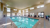 This indoor aquatic facility is designed with both function and comfort in mind. The centerpiece is a generously sized rectangular lap pool with clear turquoise water, accessible via stainless steel entry rails and steps. The pool deck is finished in neutral tile and concrete, providing ample space to move around safely.

The room features soaring double-height ceilings, with clerestory windows positioned along the upper walls on multiple sides, allowing abundant natural light to fill the space throughout the day. Large floor-to-ceiling windows and glass doors line the perimeter, offering direct views of the surrounding outdoor courtyard, landscaped grounds, and adjacent multi-story residential buildings, creating a seamless visual connection between indoors and outdoors.

Along the pool deck, multiple reclining lounge chairs paired with small wooden side tables provide comfortable relaxation areas. Tile wainscoting runs along the lower walls throughout the facility, contributing a clean and durable aesthetic. Additional amenities include restroom access and dual stainless steel drinking fountains set against tiled walls. A hot tub area is also visible at one end of the pool, rounding out this well-appointed aquatic space.