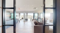 This contemporary two-floor space offers a refined blend of modern architecture and open, light-filled design. The main floor comprises the majority of the footprint and includes two distinct living areas, a hallway, and a sleek kitchen. Light hardwood flooring runs throughout, complemented by crisp white walls, recessed lighting, and clean architectural lines.

The primary living area is anchored by a striking textured feature wall with a wall-mounted television, flanked by black-framed glass partitions that allow light to flow freely between spaces. A sculptural live-edge wood bench serves as a distinctive centerpiece. Adjacent to this is a second living area bathed in natural light from tall, floor-to-ceiling windows that frame sweeping views of the surrounding landscape and skyline.

The kitchen is sleekly designed with dark cabinetry, bar-height seating, and modern appliances, offering a functional yet stylish space visible from the main living area.

The upper level opens onto a spacious covered terrace, featuring a warm wood-paneled ceiling and open-air views in multiple directions. This outdoor living area is furnished for comfort and entertaining, with panoramic vistas of rolling hills and the surrounding cityscape framed by glass railings and structural concrete columns — making it a standout feature of the entire space.
