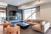 Located at 425 SW 11th St in Des Moines, Iowa, this single-floor communal space offers a thoughtfully designed open-plan layout that seamlessly integrates a fully equipped kitchen, lounge area, and multiple dining zones. Light-toned hardwood flooring runs throughout, creating a warm and cohesive aesthetic that ties the entire space together.

The kitchen serves as a central hub, featuring a large island with a white quartz countertop and bar-height seating with wishbone-style chairs. Stainless steel appliances, a built-in cooktop, double wall ovens, a full-size refrigerator, and open wooden shelving combine functionality with a clean, contemporary look. Globe pendant lights suspended from the ceiling add an inviting warmth to the area.

Adjacent to the kitchen, a comfortable lounge area is anchored by a dramatic floor-to-ceiling dark stone fireplace surround with an integrated TV. Generous seating, including a neutral sectional sofa and rounded accent chairs, creates an approachable gathering environment. Large windows allow natural light to filter in, offering views of the surrounding streetscape.

A built-in banquette dining area, upholstered in rich caramel leather against a light wood frame and teal accent wall, provides additional casual seating. Black-framed glass doors with transom windows define transitions between zones while maintaining an open, connected feel throughout this well-appointed communal space.