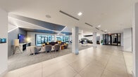Located at 3061 George Busbee Pkwy NW in Kennesaw, Georgia, this expansive single-floor amenity space is designed with a sleek, contemporary aesthetic that prioritizes both comfort and functionality. The open-plan layout is unified by a striking geometric LED lighting system, with angular lines of light traversing the ceilings and accent walls, creating a dynamic visual identity throughout.

The entrance welcomes visitors with large-format tile flooring that transitions seamlessly into the main living area. The central lounge is anchored by a generous sectional sofa, accent chairs in grey and yellow, and casual coffee tables, fostering an inviting atmosphere for relaxation and socializing. Adjacent to this, multiple high-top tables and rounded café-style seating arrangements provide flexible spaces for gathering or working.

A dedicated media lounge features plush seating, a wall-mounted flat-screen TV, and a bold geometric accent wall illuminated by recessed LED strips. A cozy booth nook with dark leather banquettes and individual tables offers a more private setting. The entertainment area includes a billiard table set beneath a recessed ceiling detail, bathed in natural light from floor-to-ceiling windows with direct access to an outdoor courtyard. A glass-partitioned fitness center is also visible, equipped with exercise equipment and bright linear lighting.