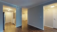 This single-floor residence offers a well-organized, open layout designed for comfortable everyday living. The interior is immediately distinguished by its cool blue-gray wall tones, which lend a calm and contemporary atmosphere throughout the space. Dark hardwood flooring runs continuously across the main living areas, providing a rich contrast against the light-colored walls and white baseboards.

Crown molding is a consistent architectural detail that adds a refined finish to the ceilings, which feature a textured surface. The open corridor-style layout connects the main living area to the entryway, creating a natural flow between spaces. The entry door is a classic paneled white design, enhancing the clean aesthetic of the unit.

The kitchen, visible from the main living area, features white cabinetry and standard appliances, offering a functional and tidy cooking space. HVAC vents are integrated into the ceiling and walls, ensuring climate comfort throughout. Light switches and electrical outlets are thoughtfully placed at standard heights.

Overall, the space presents a straightforward, move-in-ready layout with consistent finishes — making it a practical and visually cohesive residential unit suited for a variety of lifestyles.