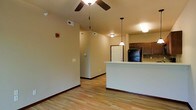 This single-floor residence offers a clean, functional layout with an open-concept design that seamlessly connects the living area to the kitchen. The living space features light hardwood-style flooring that runs throughout, complemented by warm neutral walls and dark wood baseboards that add a grounding contrast. A ceiling fan overhead provides practical comfort, while the generous ceiling height lends a sense of openness to the main living area.

The kitchen is defined by a raised breakfast bar that serves as a natural divider between the cooking and living spaces. Dark wood cabinetry provides ample storage and pairs well with the tile backsplash, while a black refrigerator anchors the kitchen's practical design. Pendant lighting hangs above the bar area, adding a touch of warmth and focused illumination to the space.

A hallway visible from the main living area leads further into the unit, suggesting additional private rooms beyond. A paneled entry door at the end of the hallway adds a classic touch. The overall ambiance is warm and welcoming, with consistent finishes and thoughtful lighting creating a cohesive, move-in-ready living environment suited for everyday comfort.