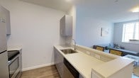 This 2-bedroom, 2-bathroom single-floor residence showcases a clean, modern aesthetic with contemporary finishes throughout. The kitchen features sleek white quartz countertops that extend along a galley-style layout, complemented by flat-front gray cabinetry with brushed metal hardware. A stainless steel undermount sink with a modern arc faucet anchors the workspace, while a built-in dishwasher adds everyday convenience. A tile backsplash adds subtle texture behind the sink area. The kitchen opens toward the living area, creating a connected, open-concept feel that encourages easy flow between spaces. Light wood-look flooring runs throughout, adding warmth and continuity to the neutral color palette of soft whites and cool grays. The living area, visible from the kitchen, provides a comfortable gathering space. The overall ambiance is bright and contemporary, with a minimalist design approach that keeps the interiors feeling fresh and uncluttered. The two bedrooms and two full bathrooms offer practical accommodation for residents seeking a well-appointed, move-in-ready space with modern conveniences.