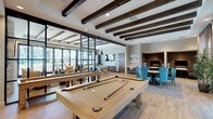 This single-floor community amenity space is thoughtfully designed to blend comfort and functionality in a modern, upscale setting. Exposed dark wood ceiling beams run throughout, adding warmth and architectural character to the bright, open interior. Wood-look flooring flows seamlessly across the space, unifying the distinct activity zones.

The entry corridor welcomes visitors through sleek, floor-to-ceiling glass and dark metal-framed doors, opening into a spacious interior. A dedicated game area features a full-size billiards table as its centerpiece, positioned beneath the striking beamed ceiling. Adjacent to this is a well-appointed kitchen with a large white quartz island, dark wood cabinetry, stainless steel appliances including a double oven, and a bold geometric tile backsplash. Industrial-style pendant lights hang above the island, adding to the contemporary aesthetic.

The lounge area offers a comfortable gathering space anchored by a floor-to-ceiling stacked stone fireplace with a mounted flat-screen television above. Plush seating in warm gold and tan tones invites relaxation. Large sliding glass doors and expansive windows flood the space with natural light and provide direct views and access to an outdoor terrace and surrounding landscape, creating a seamless indoor-outdoor connection throughout.