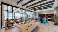 This single-floor community amenity space is thoughtfully designed to blend comfort and functionality in a modern, upscale setting. Exposed dark wood ceiling beams run throughout, adding warmth and architectural character to the bright, open interior. Wood-look flooring flows seamlessly across the space, unifying the distinct activity zones.

The entry corridor welcomes visitors through sleek, floor-to-ceiling glass and dark metal-framed doors, opening into a spacious interior. A dedicated game area features a full-size billiards table as its centerpiece, positioned beneath the striking beamed ceiling. Adjacent to this is a well-appointed kitchen with a large white quartz island, dark wood cabinetry, stainless steel appliances including a double oven, and a bold geometric tile backsplash. Industrial-style pendant lights hang above the island, adding to the contemporary aesthetic.

The lounge area offers a comfortable gathering space anchored by a floor-to-ceiling stacked stone fireplace with a mounted flat-screen television above. Plush seating in warm gold and tan tones invites relaxation. Large sliding glass doors and expansive windows flood the space with natural light and provide direct views and access to an outdoor terrace and surrounding landscape, creating a seamless indoor-outdoor connection throughout.