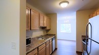 This single-floor space features a well-appointed kitchen that flows seamlessly into a dining area. The kitchen is outfitted with warm honey-toned wood cabinetry lining the upper and lower walls, providing generous storage. A granite-look laminate countertop runs the length of the kitchen, complemented by a stainless steel sink and built-in appliances, including a dishwasher and microwave. A flush-mount ceiling light illuminates the kitchen area, keeping the workspace bright and functional. Adjacent to the kitchen, the dining area benefits from a large window that invites natural light and offers a view of the surrounding greenery. An elegant chandelier hangs above, adding a refined touch to the space. The flooring transitions from the kitchen into the dining area, where light hardwood-style flooring enhances the open, airy feel. The neutral wall tones throughout create a cohesive and welcoming ambiance, making the space both practical and comfortable for everyday living.