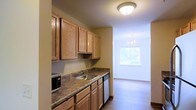 This single-floor space features a well-appointed kitchen that flows seamlessly into a dining area. The kitchen is outfitted with warm honey-toned wood cabinetry lining the upper and lower walls, providing generous storage. A granite-look laminate countertop runs the length of the kitchen, complemented by a stainless steel sink and built-in appliances, including a dishwasher and microwave. A flush-mount ceiling light illuminates the kitchen area, keeping the workspace bright and functional. Adjacent to the kitchen, the dining area benefits from a large window that invites natural light and offers a view of the surrounding greenery. An elegant chandelier hangs above, adding a refined touch to the space. The flooring transitions from the kitchen into the dining area, where light hardwood-style flooring enhances the open, airy feel. The neutral wall tones throughout create a cohesive and welcoming ambiance, making the space both practical and comfortable for everyday living.