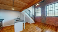 Located on Atwells Avenue in Providence's vibrant Federal Hill neighborhood, this two-floor space blends industrial character with modern finishes throughout. The main level immediately impresses with its warm tongue-and-groove wood plank ceilings supported by exposed beams, rich hardwood floors, and a dramatic exposed brick accent wall punctuated by large industrial-style windows that draw in natural light. A ceiling fan adds comfort to the open living area, which flows seamlessly into the kitchen.

The kitchen is well-appointed with white cabinetry, stainless steel appliances including a full-size refrigerator, range, and built-in microwave, complemented by granite-style countertops. A peninsula with an undermount sink and dishwasher provides additional prep space and separates the kitchen from the main living area. A sleek steel-railed staircase with wood treads connects the two floors.

The upper level features carpeted bedroom space with a wall-mounted split system for climate control and an exposed wood beam ceiling adding rustic warmth. A dedicated walk-in closet with built-in shelving provides generous storage. The bathroom includes hardwood flooring, a tub-shower combination, and clean white finishes, maintaining the cohesive aesthetic found throughout the space.