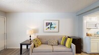 This single-floor unit offers a thoughtfully arranged layout that maximizes functionality across its living, dining, kitchen, and bathroom areas. The living room serves as the central gathering space, featuring light-toned hardwood-style flooring, soft neutral walls, and a textured ceiling. A comfortable sofa accented with colorful throw pillows anchors the room, complemented by a side table with a lamp and framed wall art that adds a touch of personality to the space.

Adjacent to the living area, the dining space makes a strong visual statement with a floor-to-ceiling stone-textured accent wall and a wall-mounted flat-screen television. A compact dining table with dark chairs is positioned near a window that allows natural light to filter in, creating an inviting atmosphere for meals.

The kitchen is equipped with white cabinetry, a built-in microwave, an electric cooktop, and generous granite-style countertops that provide ample workspace. A dishwasher and sink complete the functional setup, making the kitchen both practical and clean in appearance.

The bathroom features a single-sink vanity with a mirror, white cabinetry, and an adjacent tub-shower combination lined with square tiles. A safety grab bar is installed near the tub, and warm wood trim around the doorway adds a classic touch. Overall, the space presents a clean, functional, and comfortable environment.