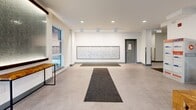 Located at 647 Goldstream Avenue in Langford, British Columbia, this ground-floor common area serves as the main entry and circulation hub of the building. The space is divided into two key zones: a welcoming lounge area and a connected hallway corridor, together offering a well-organized and functional layout.

The lounge area is tastefully appointed with a set of deep blue leather armchairs arranged around a striking live-edge wood coffee table, complemented by matching natural wood side tables. A large round decorative mirror anchors the wall above a floral arrangement, while a tall arc floor lamp adds warm ambient lighting. The entrance door provides direct access from the exterior, and a low-pile carpet runner defines the entry transition zone.

The hallway is clean and contemporary, finished with large-format light grey tile flooring and crisp white walls. Two stainless steel elevator doors are prominently positioned along one wall, accompanied by a geometric dark-panelled accent feature that adds visual interest. Recessed ceiling lighting illuminates the corridor evenly throughout.

An additional area houses a full bank of individual resident mailboxes mounted flush to the wall, providing convenient mail access. Slim industrial-style benches and wood accent pieces are placed thoughtfully along the hallway, reinforcing the modern yet warm aesthetic of the space overall.