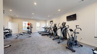 This fitness center in Clarksville, Tennessee, offers a thoughtfully designed workout space that makes efficient use of its single-floor layout. The room features crisp white walls accented by elegant crown molding, recessed lighting, and neutral carpet flooring, creating a clean and motivating environment. Multiple windows allow natural light to fill the space, adding an open, airy feel.

The cardio section is well-appointed, housing several treadmills, elliptical machines, and an air bike, each paired with mounted flat-screen televisions for entertainment during workouts. A dedicated strength training area includes a multi-function cable machine, a tiered dumbbell rack with a full range of weights, and adjustable workout benches suitable for a variety of exercises. A medicine ball rack with assorted weighted balls rounds out the functional fitness offerings.

The space is organized to allow comfortable movement between equipment zones, with adequate open floor area for stretching or bodyweight exercises. Motivational wall décor adds a personal touch to the otherwise streamlined aesthetic. The room is climate-controlled and provides easy access via clearly marked exits. Overall, this fitness center delivers a comprehensive range of exercise options within a well-maintained and welcoming facility.