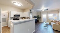 Located at 1128 Bella Springs View in Colorado Springs, Colorado, this single-story residence offers a thoughtfully designed open-concept layout that seamlessly connects its key living spaces. The kitchen is a standout feature, showcasing crisp white cabinetry, stainless steel appliances including a range and built-in microwave, a mosaic tile backsplash, and a generously sized peninsula with light stone countertops — ideal for casual dining or meal prep. Crown molding throughout adds a refined architectural touch. Rich wood-look flooring flows through the main living areas, complementing the neutral warm-toned walls. A built-in desk nook off the kitchen provides a dedicated workspace, adding practical versatility to the floor plan. The living room is bright and welcoming, with natural light pouring in through large windows fitted with blinds, and a ceiling fan overhead. A door from the living area leads to the outside. The bedroom is carpeted and serene, featuring crown molding, a ceiling fan, and a window with natural light. The bathroom offers a sleek white vanity with stone countertops and wood-look flooring, with direct access to a walk-in closet with built-in shelving, completing this cohesive and functional living space.