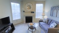 This single-floor residence offers a thoughtfully designed living area that balances comfort with timeless style. The living room serves as the heart of the space, centered around a classic white-mantled fireplace adorned with decorative accents, including ceramic vases and a striking round mirror with geometric detailing. The room is bathed in natural light, thanks to multiple large windows fitted with horizontal blinds that allow for easy light control. Neutral, warm-toned walls and plush carpeting throughout create a soft, welcoming ambiance. The furnishings are tastefully arranged, featuring a contemporary sofa in muted grey tones, a round glass-top coffee table, and coordinating accent seating. A side table with a classic lamp adds a refined touch to the corner of the room. The overall design palette is clean and understated, with subtle pops of teal and earth tones in the decorative elements that tie the space together. The layout feels open and well-proportioned, with a natural flow that makes the living area both functional and aesthetically pleasing — ideal for both relaxed everyday living and comfortable entertaining.