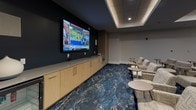 Located at 265 Communication Way in Hyannis, Massachusetts, this single-floor media lounge is designed for comfortable group viewing and social gathering. The room is anchored by a large wall-mounted flat-screen display set against a deep navy accent wall, complemented by a long run of light wood cabinetry beneath it, providing ample storage. Rows of woven fabric lounge chairs are arranged in a theater-style configuration, each accompanied by small round side tables for convenience. The flooring features a striking blue and gold patterned carpet that adds visual depth and energy to the space. A coffered ceiling with recessed lighting and integrated cove illumination creates a layered, polished ambiance throughout. Decorative framed artwork in coordinating blue and gold tones adorns one wall, reinforcing the cohesive color palette. A beverage station with a compact refrigerator and additional cabinetry is tucked along one side, enhancing the lounge's hospitality functionality. The neutral gray walls and clean white trim offer a refined contrast to the bold design elements, while integrated wall-mounted speakers support an immersive audio-visual experience. The overall design balances modern aesthetics with comfortable, functional furnishings suited for media viewing or group events.