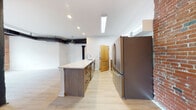This single-floor loft space masterfully combines industrial heritage with contemporary design. The open main living area is defined by striking exposed brick walls, original cast iron columns, and overhead ductwork that speak to the building's historic roots. Light hardwood flooring runs throughout, complementing the bright white walls and creating a warm, airy atmosphere. Multiple wood-framed windows flood the space with natural light while offering views of the surrounding streetscape.

The kitchen is a functional centerpiece, featuring a generous island with a stone-look countertop, dark wood cabinetry with integrated hardware, and stainless steel appliances including a large French door refrigerator. White upper cabinetry and an in-unit washer/dryer add practicality to the space. An exposed brick accent wall in the kitchen ties the room seamlessly to the industrial aesthetic found throughout.

The open living and sleeping areas benefit from the expansive footprint, with exposed beams overhead and abundant natural light streaming through the large windows set within the original brick facade. The bathroom offers a clean, modern retreat finished with large-format neutral tiles, a glass-enclosed walk-in shower, and a sleek floating vanity, providing a refined contrast to the raw industrial character of the surrounding space.