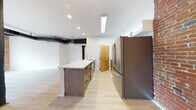 This single-floor loft space masterfully combines industrial heritage with contemporary design. The open main living area is defined by striking exposed brick walls, original cast iron columns, and overhead ductwork that speak to the building's historic roots. Light hardwood flooring runs throughout, complementing the bright white walls and creating a warm, airy atmosphere. Multiple wood-framed windows flood the space with natural light while offering views of the surrounding streetscape.

The kitchen is a functional centerpiece, featuring a generous island with a stone-look countertop, dark wood cabinetry with integrated hardware, and stainless steel appliances including a large French door refrigerator. White upper cabinetry and an in-unit washer/dryer add practicality to the space. An exposed brick accent wall in the kitchen ties the room seamlessly to the industrial aesthetic found throughout.

The open living and sleeping areas benefit from the expansive footprint, with exposed beams overhead and abundant natural light streaming through the large windows set within the original brick facade. The bathroom offers a clean, modern retreat finished with large-format neutral tiles, a glass-enclosed walk-in shower, and a sleek floating vanity, providing a refined contrast to the raw industrial character of the surrounding space.