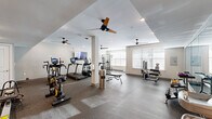 This fitness center offers a thoughtfully designed, single-floor layout that divides the space into distinct functional zones, providing a comprehensive workout experience. The main cardio and strength training area features durable rubber flooring and is outfitted with multiple treadmills, ellipticals, a stationary bike, and a cable machine, complemented by a full rack of colorful dumbbells. Ceiling fans and large windows with horizontal blinds allow natural light to fill the room while maintaining a comfortable, well-ventilated environment. A wall-mounted flat-screen TV adds an entertainment element for those engaged in cardio sessions. Connected to the main gym area is a dedicated group exercise and functional training studio, distinguished by warm hardwood-style flooring, bold navy accent walls, and a wall-to-wall mirror with an integrated ballet barre. The studio is equipped with stability balls, medicine balls, resistance bands, and other functional training tools neatly organized on racks. Ceiling fans and recessed lighting enhance the open, airy atmosphere. A private room with a glass-panel door provides additional flexibility for focused or private training sessions. Together, the zones create a versatile and inviting fitness facility suitable for a wide range of workout styles.