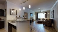 This well-appointed single-floor residence showcases an open-concept design that seamlessly connects the kitchen and living areas, creating a cohesive and inviting atmosphere. The kitchen features a curved granite-topped breakfast bar, perfect for casual dining or entertaining, complemented by a striking set of pendant lights on a track rail that adds both functionality and contemporary style. Crown molding along the ceiling adds a refined architectural touch throughout. The living area is generously proportioned, bathed in natural light streaming through large windows dressed with elegant dark curtains. Warm hardwood-style flooring extends throughout the space, lending a cohesive and welcoming feel underfoot. A ceiling fan anchors the living room, ensuring comfort year-round. The living space is versatile and accommodating, with ample room for a full seating arrangement. Framed artwork adorns the walls, contributing to the polished, move-in-ready aesthetic. The overall ambiance is warm and modern, blending practical design elements with tasteful finishes to create a comfortable and stylish everyday living environment.