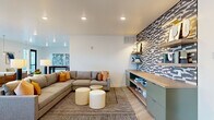 This single-floor space offers a thoughtfully designed open-plan layout that seamlessly integrates a living room, dining area, and kitchen. Light hardwood flooring runs throughout, creating a cohesive and airy feel across the entire space.

The living room serves as the centerpiece, anchored by a large sectional sofa and a bold, graphic-patterned accent wall featuring built-in wood shelving and a teal cabinetry unit below. Recessed lighting illuminates the area, complementing the clean white ceiling.

The dining area is defined by a warm built-in banquette upholstered in neutral tones, paired with round white tables and leather accent chairs. Pendant lights with gold hardware hang overhead, and a large window frames views of an outdoor terrace, flooding the space with natural light.

The kitchen is finished with warm wood cabinetry, white quartz countertops, and a bright subway-tiled backsplash. A stainless steel French door refrigerator and an under-mount sink contribute to its clean, functional aesthetic.

A separate windowed area with direct exterior door access opens to an outdoor terrace, offering additional flexibility. Throughout, the design balances modern minimalism with warm material choices, resulting in a polished and inviting atmosphere.