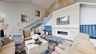 Located at 265 Communication Way in Hyannis, MA, this upscale community amenity space presents a refined, coastal-inspired design throughout. The grand lobby serves as the centerpiece, boasting soaring double-height ceilings, a striking staircase with sleek metal railings, and a floor-to-ceiling stacked stone fireplace topped with a wall-mounted TV. Plush seating arrangements and a richly patterned blue-and-neutral carpet evoke an elevated, welcoming ambiance.

An expansive lounge area features dramatic floor-to-ceiling windows that flood the space with natural light and frame outdoor views, complemented by a grand piano, woven accent chairs, and sculptural pendant lighting. The dining area accommodates multiple seating configurations on coordinating patterned carpet under a coffered ceiling with a statement chandelier, with direct access to an outdoor patio through large glass doors.

A dedicated event or meeting room offers flexible seating with rows of upholstered chairs, a small lounge vignette, and a curated gallery of large-scale abstract artwork in soothing blue and gray tones. The catering kitchen features a long countertop workspace with warm wood cabinetry, mosaic tile accents, a built-in microwave, and a wall-mounted TV, designed to support events and gatherings with ease.