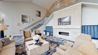 Located at 265 Communication Way in Hyannis, MA, this upscale community amenity space presents a refined, coastal-inspired design throughout. The grand lobby serves as the centerpiece, boasting soaring double-height ceilings, a striking staircase with sleek metal railings, and a floor-to-ceiling stacked stone fireplace topped with a wall-mounted TV. Plush seating arrangements and a richly patterned blue-and-neutral carpet evoke an elevated, welcoming ambiance.

An expansive lounge area features dramatic floor-to-ceiling windows that flood the space with natural light and frame outdoor views, complemented by a grand piano, woven accent chairs, and sculptural pendant lighting. The dining area accommodates multiple seating configurations on coordinating patterned carpet under a coffered ceiling with a statement chandelier, with direct access to an outdoor patio through large glass doors.

A dedicated event or meeting room offers flexible seating with rows of upholstered chairs, a small lounge vignette, and a curated gallery of large-scale abstract artwork in soothing blue and gray tones. The catering kitchen features a long countertop workspace with warm wood cabinetry, mosaic tile accents, a built-in microwave, and a wall-mounted TV, designed to support events and gatherings with ease.