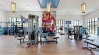 Located at 5400 Broken Sound Blvd NW in Boca Raton, FL, this single-story fitness center offers a well-appointed workout environment designed to inspire and accommodate a comprehensive exercise routine. The space features soaring ceilings that lend an open, airy feel, complemented by large, multi-pane black-framed windows that flood the interior with natural light and provide views of the surrounding Florida greenery.

The design aesthetic is cohesive and polished, with warm wood-look flooring throughout, crisp white upper walls, and distinctive blue wainscoting that adds a refined, upscale character to the space. Elegant gold pendant light fixtures hang from the ceiling, enhancing the upscale ambiance.

The fitness center is comprehensively equipped with a full cable/multi-functional strength training system, a dedicated dumbbell rack with mirror, rowing machines, treadmills, ellipticals, and additional resistance equipment. Multiple wall-mounted flat-screen televisions are positioned throughout for entertainment during workouts. A motivational large-scale fitness mural serves as a striking focal point along one wall.

Additional amenities include built-in storage shelving, a compact refrigerator, and blue double doors providing access to storage or additional facilities. The space benefits from direct outdoor access through glass doors, connecting the interior to the exterior grounds.