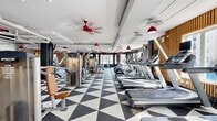 Located at 113 Ellington Blvd in Gaithersburg, MD, this well-appointed fitness center offers a generously sized, single-floor exercise space designed to accommodate a comprehensive range of workout activities. The interior is immediately striking thanks to its bold black-and-white geometric triangle-patterned flooring, which creates a dynamic visual contrast against the bright white exposed ceiling with visible ductwork and piping — a nod to modern industrial design.

Distinctive red pendant lights and matching red ceiling fans punctuate the space overhead, adding a vibrant pop of color and ensuring consistent airflow throughout. Warm wood slat accent walls provide a natural, textural counterpoint to the darker charcoal feature walls, contributing to a balanced and energetic ambiance.

The exercise floor is comprehensively equipped with a full row of treadmills, ellipticals, and cardio machines, as well as a wide array of Precor strength-training machines, cable systems, free weights, benches, and cycling bikes. Large windows along the perimeter flood the space with abundant natural light and offer views of the surrounding streetscape, enhancing the openness of the environment. A mirrored wall section supports strength and stretching areas. The layout is thoughtfully organized to provide clear zones for both cardio and strength training.