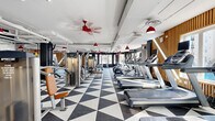 Located at 113 Ellington Blvd in Gaithersburg, MD, this well-appointed fitness center offers a generously sized, single-floor exercise space designed to accommodate a comprehensive range of workout activities. The interior is immediately striking thanks to its bold black-and-white geometric triangle-patterned flooring, which creates a dynamic visual contrast against the bright white exposed ceiling with visible ductwork and piping — a nod to modern industrial design.

Distinctive red pendant lights and matching red ceiling fans punctuate the space overhead, adding a vibrant pop of color and ensuring consistent airflow throughout. Warm wood slat accent walls provide a natural, textural counterpoint to the darker charcoal feature walls, contributing to a balanced and energetic ambiance.

The exercise floor is comprehensively equipped with a full row of treadmills, ellipticals, and cardio machines, as well as a wide array of Precor strength-training machines, cable systems, free weights, benches, and cycling bikes. Large windows along the perimeter flood the space with abundant natural light and offer views of the surrounding streetscape, enhancing the openness of the environment. A mirrored wall section supports strength and stretching areas. The layout is thoughtfully organized to provide clear zones for both cardio and strength training.