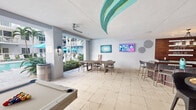 This single-level communal lounge and recreation space is designed to encourage social interaction and leisure. The most striking architectural feature is the ceiling, which showcases a sweeping teal and gold sculptural element that adds a bold, resort-inspired focal point to the room. Light-toned stone tile flooring runs throughout, contributing to a bright, clean aesthetic that complements the crisp white walls.

The space is organized to accommodate a variety of activities. A pool or shuffleboard table anchors one end of the room, while a central dining-height table surrounded by modern bucket-style chairs provides a casual gathering area. A bar-height counter with tall stools along one wall offers additional seating, ideal for socializing or watching the wall-mounted flat-screen television nearby. Colorful contemporary artwork decorates the walls, enhancing the lively atmosphere.

A wide open passage connects the interior directly to an outdoor pool area, where lush greenery and teal patio umbrellas are visible, allowing abundant natural light to flood the space. A ceiling fan aids in airflow. Wall-mounted sconce lighting completes the amenity-focused design, creating a welcoming, energetic environment suited for both relaxation and entertainment.