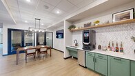 Located at 14211 Akron Ave in Rosemount, MN, this single-floor community lounge is a well-appointed space designed to foster both social interaction and focused gathering. The layout flows seamlessly across several distinct zones, each serving a specific purpose while contributing to a cohesive, modern aesthetic.

A standout kitchenette anchors one side of the space, featuring sage green cabinetry, a white quartz countertop, a built-in sink, an under-counter refrigerator, and a commercial-grade coffee machine. Open wood shelving above the counter adds a warm, decorative touch with curated plants and accents. Adjacent to the kitchenette is a dining area centered around a solid wood table with mid-century style chairs, illuminated by an industrial-modern geometric pendant light. A wall-mounted flat-screen television is positioned nearby for presentations or shared viewing.

A built-in banquette runs along a deep charcoal accent wall, paired with small pedestal tables and bistro-style chairs, creating an inviting lounge setting accented by framed botanical and geometric artwork. A glass-partitioned private room with black-framed doors offers a quieter enclave for meetings or focused work. Light wood-look flooring, a drop tile ceiling with recessed lighting, and natural light from exterior-facing doors complete the bright, clean, and welcoming ambiance of this versatile space.