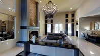 This single-story space makes a striking impression through its dramatic design elements and upscale aesthetic. The centerpiece of the main lounge area is a floor-to-ceiling stacked stone fireplace, which anchors the room with a bold, textural presence. Above, an ornate wrought-iron and crystal chandelier cascades from the soaring ceiling, casting a warm, ambient glow throughout the space.

The exceptionally high ceilings amplify the sense of grandeur, while tall windows and glass doors at the far end of the room invite natural light and offer views of the exterior. Dark curtain panels flank the windows, adding a sense of depth and formality to the room.

At the heart of the lounge sits a large, sectional sofa in rich charcoal upholstery, arranged around a sleek, dark-surface coffee table. Decorative accents, including sculptural vases and reflective objects, contribute to the polished, contemporary ambiance. The light-toned floors contrast elegantly with the dark furnishings, enhancing the room's sophisticated character. Overall, this space exudes a refined, modern atmosphere well-suited for upscale social or professional gatherings.