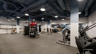 Located at 1133 W Lawrence Ave in Chicago's Uptown neighborhood, this fitness center offers a comprehensive, well-organized workout facility across a single floor. Guests are welcomed through a dramatic entrance hallway lined floor-to-ceiling with bold blue-and-white tropical botanical wallpaper, setting a distinctive visual tone against polished concrete floors and an exposed industrial ceiling with painted ductwork and piping.

The main workout area is expansive and open, featuring rubber flooring throughout for comfort and safety. It is divided into functional zones catering to a wide range of fitness activities. The strength training section is equipped with cable machines, a multi-functional rack, free weights including dumbbells and barbells, adjustable benches, and a punching bag. Full-length mirrors line portions of the walls, enhancing the sense of space and supporting form-focused training.

A dedicated cardio section houses treadmills, ellipticals, stationary bikes, and rowing machines. A raised mezzanine-style platform area with metal railings and stair access provides additional space for equipment and training activities.

The facility is illuminated by industrial pendant and round flush-mount lights, maintaining a bright yet utilitarian atmosphere. Accent walls featuring the same tropical wallpaper motif appear throughout, adding personality to the otherwise industrial aesthetic. A framed coastal artwork provides a calm visual focal point within the dynamic training environment.
