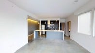 This single-floor space showcases a contemporary open-plan design centered around a well-appointed kitchen and living area. The polished concrete floors run throughout, lending an industrial yet refined aesthetic that complements the clean white walls and ceilings fitted with recessed lighting.

The kitchen is a standout feature, outfitted with dark upper and lower cabinetry contrasted by a black subway tile backsplash. Stainless steel appliances, including a refrigerator, range, and microwave, provide a professional-grade feel. A large kitchen island with a light-toned countertop serves as a natural focal point, offering additional workspace and seating potential. Pendant lights hang above the island, adding warmth to the otherwise neutral palette.

An accent wall with integrated ambient LED strip lighting adds a warm, golden glow to one section of the living area, creating visual depth and dimension. The open layout allows for flexible furniture arrangements, while a corridor visible in the background leads to additional rooms. The overall ambiance is clean, modern, and functional, ideal for those who appreciate minimalist, urban design.