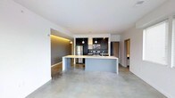 This single-floor space showcases a contemporary open-plan design centered around a well-appointed kitchen and living area. The polished concrete floors run throughout, lending an industrial yet refined aesthetic that complements the clean white walls and ceilings fitted with recessed lighting.

The kitchen is a standout feature, outfitted with dark upper and lower cabinetry contrasted by a black subway tile backsplash. Stainless steel appliances, including a refrigerator, range, and microwave, provide a professional-grade feel. A large kitchen island with a light-toned countertop serves as a natural focal point, offering additional workspace and seating potential. Pendant lights hang above the island, adding warmth to the otherwise neutral palette.

An accent wall with integrated ambient LED strip lighting adds a warm, golden glow to one section of the living area, creating visual depth and dimension. The open layout allows for flexible furniture arrangements, while a corridor visible in the background leads to additional rooms. The overall ambiance is clean, modern, and functional, ideal for those who appreciate minimalist, urban design.