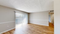 This efficiently designed single-story home offers three bedrooms, one bathroom, and a combined kitchen and living area, all connected by three hallways. Light wood flooring flows consistently throughout the entire space, complementing the neutral greige wall tones and crisp white trim and baseboards that appear in every room.

The living area is a standout, featuring a textured ceiling, a decorative chair rail, and a full-width sliding glass door fitted with vertical blinds that opens to an outdoor view, allowing generous natural light to fill the room. An entry door with a chair rail detail opens into this same space.

The kitchen is well-appointed with white upper cabinetry, a stainless steel sink, a dishwasher, a range, and a refrigerator, all set against a wood floor and illuminated by a recessed ceiling light panel.

The primary bedroom is the most spacious of the three, featuring a closet and a door that connects to the hallway. The two additional bedrooms each include a window with views to the outdoors, providing ample natural light and ventilation. All bedrooms share the same cohesive flooring and wall finishes.

The bathroom is clean and functional, featuring a full tub-shower combination with white tile surround, a toilet, and a vanity, all finished in a bright, neutral palette.