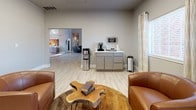 This single-floor professional office space combines functionality with a warm, inviting atmosphere styled with a distinct Western-inspired aesthetic. Light wood-look flooring runs throughout, complementing the neutral gray walls and white trim that give the space a clean, cohesive look.

The reception and waiting area serves as the heart of the space, furnished with rich cognac leather swivel chairs arranged around a striking live-edge wood coffee table atop a cowhide-style area rug. A refreshment station with gray cabinetry, a countertop coffee setup, and a water dispenser adds a welcoming, client-friendly touch. Sheer curtain panels frame the windows, allowing natural light to filter through while maintaining a comfortable ambiance.

Curated wall art featuring bold sunset landscapes — including windmills, silhouetted trees, and a lone rider on horseback — adds character and a sense of place to both the waiting and workspace areas. The workstation area is well-equipped with a centrally positioned desk island accommodating multiple workstations, a printer, and computer monitors, supported by ergonomic office chairs.

Recessed and panel ceiling lighting ensures consistent illumination throughout. The open flow between rooms enhances accessibility and creates a spacious, professional environment that feels both functional and thoughtfully designed.