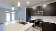 This single-floor space showcases a thoughtfully designed, modern kitchen that serves as a central highlight. The kitchen features dark espresso-toned cabinetry that extends to the ceiling, providing generous storage while creating a bold visual contrast against the crisp white quartz countertops. A large undermount sink is positioned on the kitchen island, paired with a professional-style coiled stainless steel faucet, adding both function and a sleek aesthetic.

Stainless steel appliances, including a freestanding range and an over-the-range microwave, complement the contemporary design. A textured white tile backsplash adds subtle visual interest behind the cooking area. Pendant lighting hangs above, contributing warmth and style to the space.

The light blue-gray walls create a calm, airy atmosphere throughout, while a ceiling fan ensures comfortable airflow. Natural light filters in through a window, brightening the open layout. Light-toned flooring runs throughout, enhancing the sense of openness. The overall design balances modern sophistication with practical functionality, making the kitchen both an inviting and highly usable centerpiece of the space.