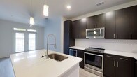 This single-floor space showcases a thoughtfully designed, modern kitchen that serves as a central highlight. The kitchen features dark espresso-toned cabinetry that extends to the ceiling, providing generous storage while creating a bold visual contrast against the crisp white quartz countertops. A large undermount sink is positioned on the kitchen island, paired with a professional-style coiled stainless steel faucet, adding both function and a sleek aesthetic.

Stainless steel appliances, including a freestanding range and an over-the-range microwave, complement the contemporary design. A textured white tile backsplash adds subtle visual interest behind the cooking area. Pendant lighting hangs above, contributing warmth and style to the space.

The light blue-gray walls create a calm, airy atmosphere throughout, while a ceiling fan ensures comfortable airflow. Natural light filters in through a window, brightening the open layout. Light-toned flooring runs throughout, enhancing the sense of openness. The overall design balances modern sophistication with practical functionality, making the kitchen both an inviting and highly usable centerpiece of the space.