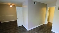 This two-floor space presents a clean, straightforward interior characterized by dark hardwood-style flooring that runs throughout the visible areas, offering a warm contrast against the light, neutral-toned walls. The walls are finished in a soft gray-beige palette, creating a calm and versatile backdrop suitable for a variety of uses. White baseboards provide a crisp border along the flooring, adding a subtle decorative detail that ties the space together. A classic six-panel white interior door is visible, consistent with the clean, traditional aesthetic carried throughout. Ceiling-mounted HVAC vents are positioned across the space, indicating a central air system for climate control. A ceiling fan is partially visible, contributing to air circulation within the room. The layout suggests a defined division of space, with a partial wall creating distinct areas within the open floor plan. The overall ambiance is neutral and move-in ready, offering a blank canvas that can be adapted to various functional arrangements. With two floors, the property provides ample vertical space and the potential for well-separated living and private areas across its levels.