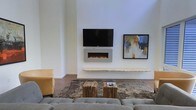 This single-floor space showcases a thoughtfully designed interior with a strong emphasis on modern, minimalist aesthetics. The living area serves as the focal point, featuring crisp white walls that create a bright and open atmosphere. A wall-mounted electric fireplace anchors the room, positioned beneath a flat-screen television and complemented by a floating live-edge wood shelf that adds a natural, organic contrast to the otherwise clean-lined design. The seating arrangement includes a plush, tufted sofa and an accent chair in warm mustard yellow, centered around a textured area rug in earthy tones. A pair of rustic wooden block coffee tables adds an artisanal touch, grounding the space with warmth and character. A framed abstract painting in bold blues and yellows adorns the wall, introducing a curated artistic element. The flooring consists of rich hardwood that runs throughout, enhancing the warmth and cohesion of the space. High, angled ceilings amplify the sense of openness, while the overall layout is both functional and visually balanced. The combination of contemporary fixtures, natural materials, and carefully selected décor makes this space an elegant and comfortable environment.