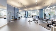 Located at 1 Harbor Point Rd in Stamford, Connecticut, this expansive fitness center offers a comprehensive, well-appointed space designed for a full range of exercise disciplines. The facility spans a single floor with a generous footprint, providing ample room for movement throughout every zone.

The main gym floor is lined with floor-to-ceiling windows that flood the space with natural light and frame views of the surrounding Harbor Point urban landscape. Light blue accent walls and durable rubber flooring give the space a clean, modern aesthetic. This area is equipped with a full array of cardio machines—including treadmills, ellipticals, stationary bikes, and rowing machines—as well as strength training stations, cable machines, free weights, benches, and squat racks with plate storage.

A dedicated studio room features a full mirrored wall, a ballet barre, and yoga mats, creating an ideal environment for barre, yoga, or stretching routines. A secondary functional training room provides foam rollers, medicine balls, stability balls, step platforms, and kettlebells for group fitness or solo conditioning work. Frosted glass partitions and large glass walls connect areas while maintaining a sense of openness. Linear overhead lighting supplements the abundant natural light throughout, ensuring a bright and energizing workout environment at any hour.