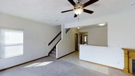 This two-story home offers a thoughtfully designed layout that balances open living spaces with practical functionality. The main level centers around a spacious living area finished with plush carpeting and light tile flooring, creating a comfortable and welcoming atmosphere. A ceiling fan with a built-in light fixture is mounted overhead, promoting air circulation and adding a classic touch to the room.

A staircase with dark wood railings rises gracefully from the main living area, serving as a central architectural feature that connects the two floors. Adjacent to the living space, a kitchen counter or breakfast bar is partially visible, suggesting an open-concept flow between the living and kitchen areas. The kitchen area appears to transition smoothly from the tiled entryway, offering a practical and accessible cooking space.

Natural light filters in through a window near the front entry, brightening the main level and enhancing the home's airy feel. The neutral color palette throughout — from the light walls to the soft flooring — provides a versatile backdrop suited to a variety of interior styles. The upper floor, accessible via the interior staircase, extends the home's living capacity, offering additional private spaces above the main level.