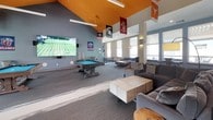 This energetic single-floor community clubhouse is designed with a lively collegiate spirit, immediately apparent through the numerous college pennants and banners suspended from the striking vaulted ceilings finished in a bold orange. The soaring ceiling height creates an airy, open atmosphere throughout the space, amplified by an abundance of natural light streaming in from large windows along multiple walls.

The main lounge area is generously appointed with plush, oversized sectional sofas, accent chairs, and modular coffee tables, offering ample seating for socializing or relaxing. A bar-height communal table with metal stools provides an additional gathering spot, ideal for casual conversations or working. An interior balcony-style overlook with metal railing adds architectural depth and visual interest, connecting different zones within the space.

The entertainment zone is a standout feature, anchored by a large wall-mounted flat-screen television, a full-size pool table, and arcade gaming machines—making it a prime destination for recreation. Rustic barn-style sliding doors and a mix of industrial and contemporary furnishings contribute to the space's distinctive character. Decorative greenery and thoughtful lighting complete the welcoming, dynamic ambiance throughout.