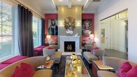 This sophisticated single-floor space combines bold design sensibilities with functional versatility. The interior is anchored by a striking lounge area centered around a marble fireplace flanked by a gold mosaic tile surround, an ornate sunburst mirror, and large-scale portrait artwork set against vivid fuchsia accent walls. Coffered ceilings add architectural depth and a sense of refinement throughout the space.

The lounge is furnished with nailhead-trim upholstered armchairs and sofas in neutral tones, accented by fuchsia throw pillows and coordinating ottomans. Decorative gold sphere accents and mirrored coffee tables add a glamorous, eclectic touch. A patterned dark carpet grounds the seating areas with visual texture.

Adjacent to the lounge is a dining area featuring a warm wood conference-style table surrounded by upholstered chairs, ideal for meetings or group gatherings. Sunburst mirrors and metallic decorative objects adorn the walls nearby, maintaining design continuity.

Large windows dressed with charcoal floor-length drapes and horizontal blinds line the perimeter, flooding the space with natural light and offering views of lush outdoor greenery. Glass-paneled doorways connect the main room to additional areas, including what appears to be a casual café or bar space with bar-height seating and tiled flooring, enhancing the overall functionality of this beautifully appointed communal environment.
