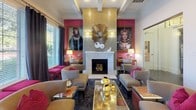 This sophisticated single-floor space combines bold design sensibilities with functional versatility. The interior is anchored by a striking lounge area centered around a marble fireplace flanked by a gold mosaic tile surround, an ornate sunburst mirror, and large-scale portrait artwork set against vivid fuchsia accent walls. Coffered ceilings add architectural depth and a sense of refinement throughout the space.

The lounge is furnished with nailhead-trim upholstered armchairs and sofas in neutral tones, accented by fuchsia throw pillows and coordinating ottomans. Decorative gold sphere accents and mirrored coffee tables add a glamorous, eclectic touch. A patterned dark carpet grounds the seating areas with visual texture.

Adjacent to the lounge is a dining area featuring a warm wood conference-style table surrounded by upholstered chairs, ideal for meetings or group gatherings. Sunburst mirrors and metallic decorative objects adorn the walls nearby, maintaining design continuity.

Large windows dressed with charcoal floor-length drapes and horizontal blinds line the perimeter, flooding the space with natural light and offering views of lush outdoor greenery. Glass-paneled doorways connect the main room to additional areas, including what appears to be a casual café or bar space with bar-height seating and tiled flooring, enhancing the overall functionality of this beautifully appointed communal environment.